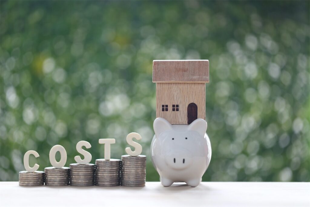 Image of rising cost in home loan depicted by stacks of growing coins and a wooden block house on top of a piggy bank